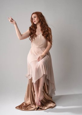 Uzun, esmer saçlı, kremsi pembe bir elbise giyen güzel bir kadın modelin tam boy portresi. Beyaz stüdyo arka planında izole edilmiş zarif oturma pozu.