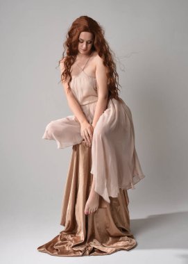 Uzun, esmer saçlı, kremsi pembe bir elbise giyen güzel bir kadın modelin tam boy portresi. Beyaz stüdyo arka planında izole edilmiş zarif oturma pozu.