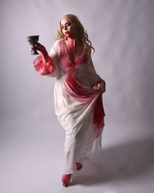  Korkunç vampir zombi gelinin portresi. Üzerinde kanlı kırmızı boya lekeleri olan şık bir Cadılar Bayramı fantazisi kostümü var. Elinde şarap kadehi var. Stüdyo arka planında izole edilmiş 