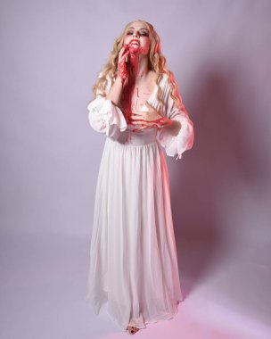  Korkunç zombi gelinin uzun bir portresi. Cadılar bayramı fantezi elbisesi ve kanlı kırmızı boya sıçraması. Ayakta durma pozu. Beyaz stüdyo arka planında izole edilmiş. 