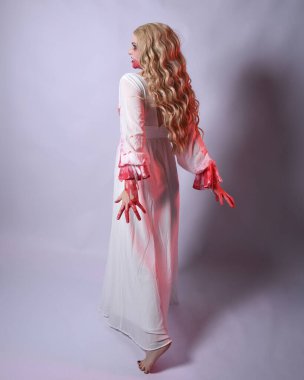  Korkunç zombi gelinin uzun bir portresi. Cadılar bayramı fantezi elbisesi ve kanlı kırmızı boya sıçraması. Ayakta durma pozu. Beyaz stüdyo arka planında izole edilmiş. 