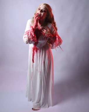  Korkunç zombi gelinin uzun bir portresi. Cadılar bayramı fantezi elbisesi ve kanlı kırmızı boya sıçraması. Ayakta durma pozu. Beyaz stüdyo arka planında izole edilmiş. 