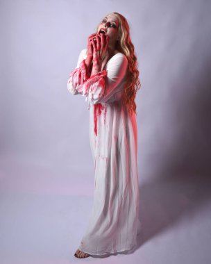  Korkunç zombi gelinin uzun bir portresi. Cadılar bayramı fantezi elbisesi ve kanlı kırmızı boya sıçraması. Ayakta durma pozu. Beyaz stüdyo arka planında izole edilmiş. 