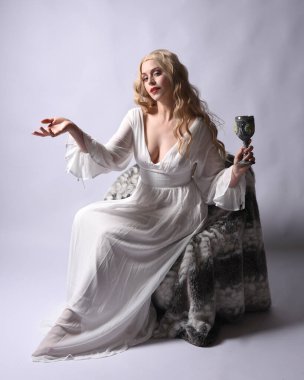Beyaz tarihi gelinlik giymiş sarışın bir kadının uzun bir portresi. Oturmuş, hareketli eller uzanıyor, stüdyo arka planında izole edilmiş..