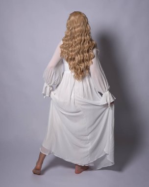 Beyaz tarihi gelinlik giymiş sarışın bir kadının uzun bir portresi. Duruş, ters dönük kameradan uzaklaşma, stüdyo arka planında izole edilmiş..