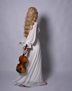 Beyaz tarihi gelinlik giymiş sarışın bir kadının uzun bir portresi. Duruş, ters dönük kameradan uzaklaşma, stüdyo arka planında izole edilmiş..