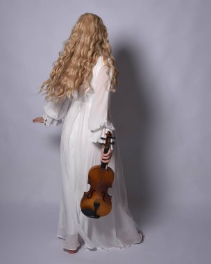 Beyaz tarihi gelinlik giymiş sarışın bir kadının uzun bir portresi. Duruş, ters dönük kameradan uzaklaşma, stüdyo arka planında izole edilmiş..