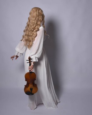 Beyaz tarihi gelinlik giymiş sarışın bir kadının uzun bir portresi. Duruş, ters dönük kameradan uzaklaşma, stüdyo arka planında izole edilmiş..