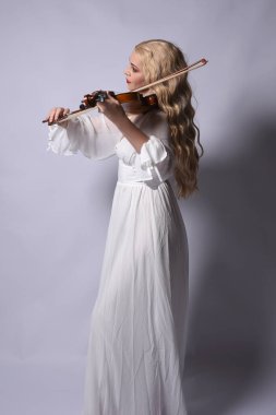Zarif beyaz cadılar bayramı elbisesi giyen güzel sarışın mankenin yakın plan portresi. Tarihi bir fantezi karakteri. Stüdyonun arka planında izole edilmiş bir keman enstrümanı tutuyor..