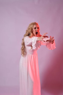 Zarif beyaz cadılar bayramı elbisesi giyen güzel sarışın mankenin yakın portresi. Aç vampir gibi uzanan kollar. Kırmızı karamsar ışıklandırma ile stüdyo arka planında izole.