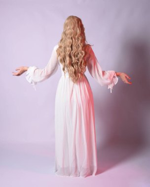 Beyaz tarihi gelinlik giymiş sarışın bir kadının uzun bir portresi. Kameradan geriye doğru yürürken poz vermek. Stüdyo arka planında izole.