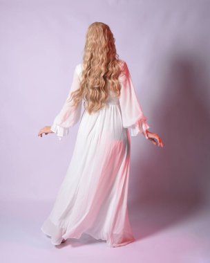 Beyaz tarihi gelinlik giymiş sarışın bir kadının uzun bir portresi. Kameradan geriye doğru yürürken poz vermek. Stüdyo arka planında izole.