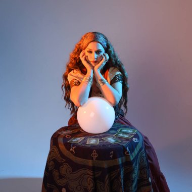 Ortaçağ fantezi falcı kostümü giyen güzel kızıl saçlı kadının yakın plan portresi seans masasında geleceği okuyan kristal küreye bakıyor. Stüdyo arka planında karamsar sinematik ışıklandırmayla izole edilmiş.