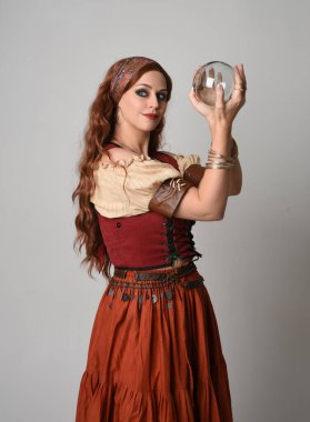 Ortaçağ bakiresi giymiş güzel kızıl saçlı bir kadının portresi. Falcı kostümü. Kristal küreyi tutarken poz vermek. Stüdyo arka planında izole.
