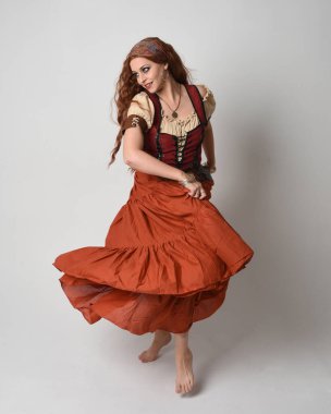 Ortaçağ bakiresi, falcı kostümü giymiş güzel kızıl saçlı bir kadının tam boy portresi. Dans hareketleriyle ayakta durmak, etek sallamak. Stüdyo arka planında izole.
