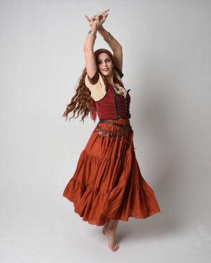 Ortaçağ bakiresi, falcı kostümü giymiş güzel kızıl saçlı bir kadının tam boy portresi. Dans hareketleriyle ayakta durmak, etek sallamak. Stüdyo arka planında izole.