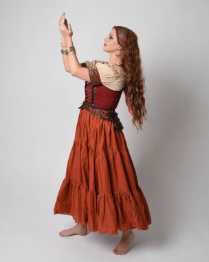 Ortaçağ bakiresi, falcı kostümü giymiş güzel kızıl saçlı bir kadının tam boy portresi. Dans hareketleriyle ayakta durmak, etek sallamak. Stüdyo arka planında izole.