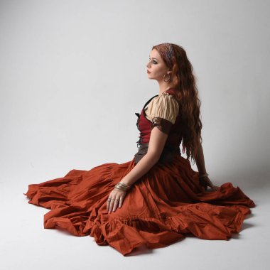 Ortaçağ bakiresi, falcı kostümü giymiş güzel kızıl saçlı bir kadının tam boy portresi. Diz çök, yere otur. Stüdyo arka planında izole.