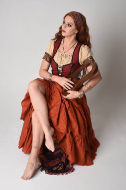 Ortaçağ bakiresi, falcı kostümü giymiş güzel kızıl saçlı bir kadının tam boy portresi. Oturma pozu, müzik aleti. Stüdyo arka planında izole.