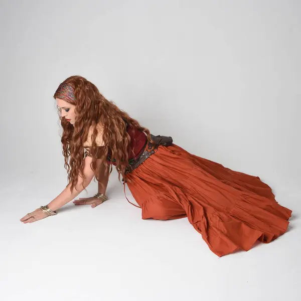Stock image Full length portrait of beautiful red haired woman wearing a medieval maiden, fortune teller costume.  Kneeling pose, sitting down on floor. isolated on studio background.