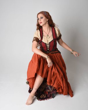 Ortaçağ bakiresi, falcı kostümü giymiş güzel kızıl saçlı bir kadının tam boy portresi. Oturma pozu, hareketli eller uzanıyor. Stüdyoda izole 