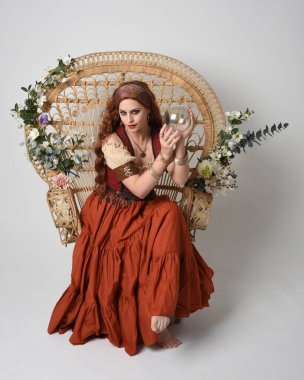 Ortaçağ bakiresi, falcı kostümü giymiş güzel kızıl saçlı bir kadının tam boy portresi. Oturma pozisyonu, elinde kristal bir küre. Stüdyo arka planında izole.