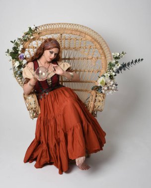 Ortaçağ bakiresi, falcı kostümü giymiş güzel kızıl saçlı bir kadının tam boy portresi. Oturma pozisyonu, elinde kristal bir küre. Stüdyo arka planında izole.