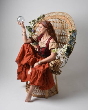 Ortaçağ bakiresi, falcı kostümü giymiş güzel kızıl saçlı bir kadının tam boy portresi. Oturma pozisyonu, elinde kristal bir küre. Stüdyo arka planında izole.