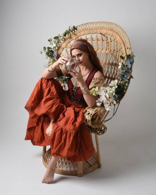 Ortaçağ bakiresi, falcı kostümü giymiş güzel kızıl saçlı bir kadının tam boy portresi. Oturma pozisyonu, elinde kristal bir küre. Stüdyo arka planında izole.