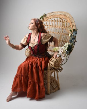 Full length portrait of beautiful red haired woman wearing a medieval maiden, fortune teller costume.  Sitting pose, with gestural hands reaching out. isolated on studio background. clipart