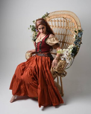 Ortaçağ bakiresi, falcı kostümü giymiş güzel kızıl saçlı bir kadının tam boy portresi. Oturma pozu, hareketli eller uzanıyor. Stüdyo arka planında izole.