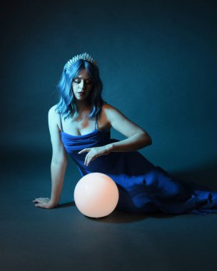 Parlak bir lamba tutan, kristal taçlı göz kamaştırıcı fantezi balo elbisesi giyen güzel, mavi saçlı kadın modelinin tam boy portresi. Oturma pozisyonu, karanlık stüdyo arka planında izole.