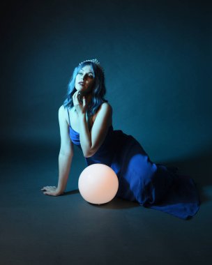 Parlak bir lamba tutan, kristal taçlı göz kamaştırıcı fantezi balo elbisesi giyen güzel, mavi saçlı kadın modelinin tam boy portresi. Oturma pozisyonu, karanlık stüdyo arka planında izole.