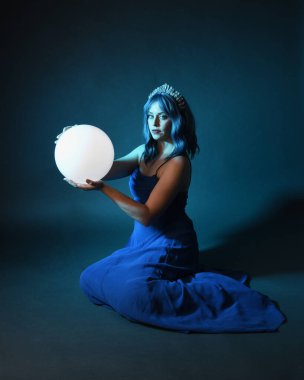 Parlak bir lamba tutan, kristal taçlı göz kamaştırıcı fantezi balo elbisesi giyen güzel, mavi saçlı kadın modelinin tam boy portresi. Oturma pozisyonu, karanlık stüdyo arka planında izole.