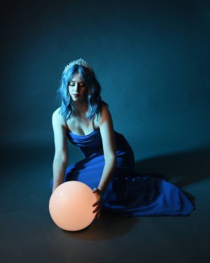 Parlak bir lamba tutan, kristal taçlı göz kamaştırıcı fantezi balo elbisesi giyen güzel, mavi saçlı kadın modelinin tam boy portresi. Oturma pozisyonu, karanlık stüdyo arka planında izole.