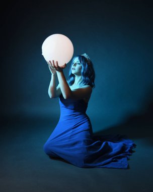 Parlak bir lamba tutan, kristal taçlı göz kamaştırıcı fantezi balo elbisesi giyen güzel, mavi saçlı kadın modelinin tam boy portresi. Oturma pozisyonu, karanlık stüdyo arka planında izole.