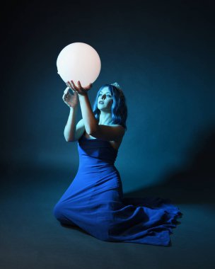 Parlak bir lamba tutan, kristal taçlı göz kamaştırıcı fantezi balo elbisesi giyen güzel, mavi saçlı kadın modelinin tam boy portresi. Oturma pozisyonu, karanlık stüdyo arka planında izole.