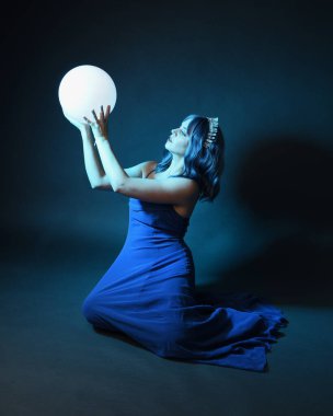 Parlak bir lamba tutan, kristal taçlı göz kamaştırıcı fantezi balo elbisesi giyen güzel, mavi saçlı kadın modelinin tam boy portresi. Oturma pozisyonu, karanlık stüdyo arka planında izole.