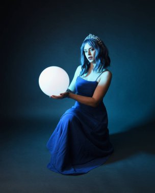 Parlak bir lamba tutan, kristal taçlı göz kamaştırıcı fantezi balo elbisesi giyen güzel, mavi saçlı kadın modelinin tam boy portresi. Oturma pozisyonu, karanlık stüdyo arka planında izole.