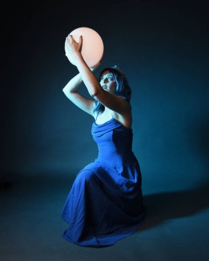 Parlak bir lamba tutan, kristal taçlı göz kamaştırıcı fantezi balo elbisesi giyen güzel, mavi saçlı kadın modelinin tam boy portresi. Oturma pozisyonu, karanlık stüdyo arka planında izole.