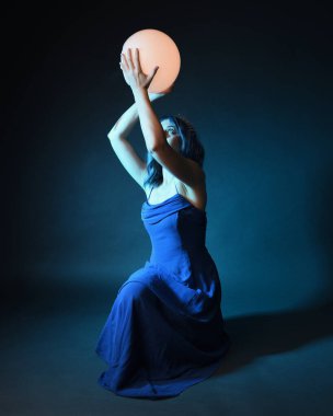 Parlak bir lamba tutan, kristal taçlı göz kamaştırıcı fantezi balo elbisesi giyen güzel, mavi saçlı kadın modelinin tam boy portresi. Oturma pozisyonu, karanlık stüdyo arka planında izole.