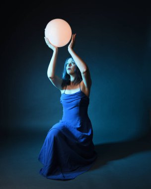 Parlak bir lamba tutan, kristal taçlı göz kamaştırıcı fantezi balo elbisesi giyen güzel, mavi saçlı kadın modelinin tam boy portresi. Oturma pozisyonu, karanlık stüdyo arka planında izole.