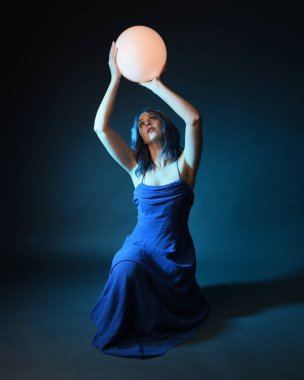 Parlak bir lamba tutan, kristal taçlı göz kamaştırıcı fantezi balo elbisesi giyen güzel, mavi saçlı kadın modelinin tam boy portresi. Oturma pozisyonu, karanlık stüdyo arka planında izole.