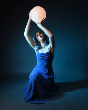 Parlak bir lamba tutan, kristal taçlı göz kamaştırıcı fantezi balo elbisesi giyen güzel, mavi saçlı kadın modelinin tam boy portresi. Oturma pozisyonu, karanlık stüdyo arka planında izole.