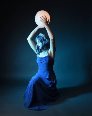 Parlak bir lamba tutan, kristal taçlı göz kamaştırıcı fantezi balo elbisesi giyen güzel, mavi saçlı kadın modelinin tam boy portresi. Oturma pozisyonu, karanlık stüdyo arka planında izole.