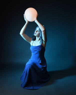 Parlak bir lamba tutan, kristal taçlı göz kamaştırıcı fantezi balo elbisesi giyen güzel, mavi saçlı kadın modelinin tam boy portresi. Oturma pozisyonu, karanlık stüdyo arka planında izole.