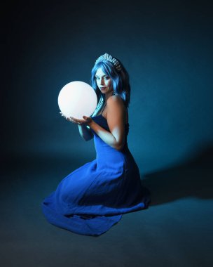 Parlak bir lamba tutan, kristal taçlı göz kamaştırıcı fantezi balo elbisesi giyen güzel, mavi saçlı kadın modelinin tam boy portresi. Oturma pozisyonu, karanlık stüdyo arka planında izole.