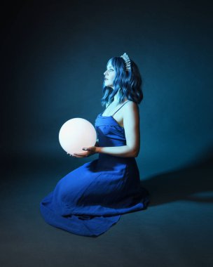 Parlak bir lamba tutan, kristal taçlı göz kamaştırıcı fantezi balo elbisesi giyen güzel, mavi saçlı kadın modelinin tam boy portresi. Oturma pozisyonu, karanlık stüdyo arka planında izole.