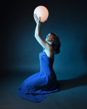 Parlak bir lamba tutan, kristal taçlı göz kamaştırıcı fantezi balo elbisesi giyen güzel, mavi saçlı kadın modelinin tam boy portresi. Oturma pozisyonu, karanlık stüdyo arka planında izole.