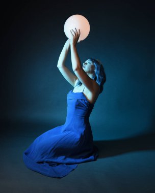 Parlak bir lamba tutan, kristal taçlı göz kamaştırıcı fantezi balo elbisesi giyen güzel, mavi saçlı kadın modelinin tam boy portresi. Oturma pozisyonu, karanlık stüdyo arka planında izole.